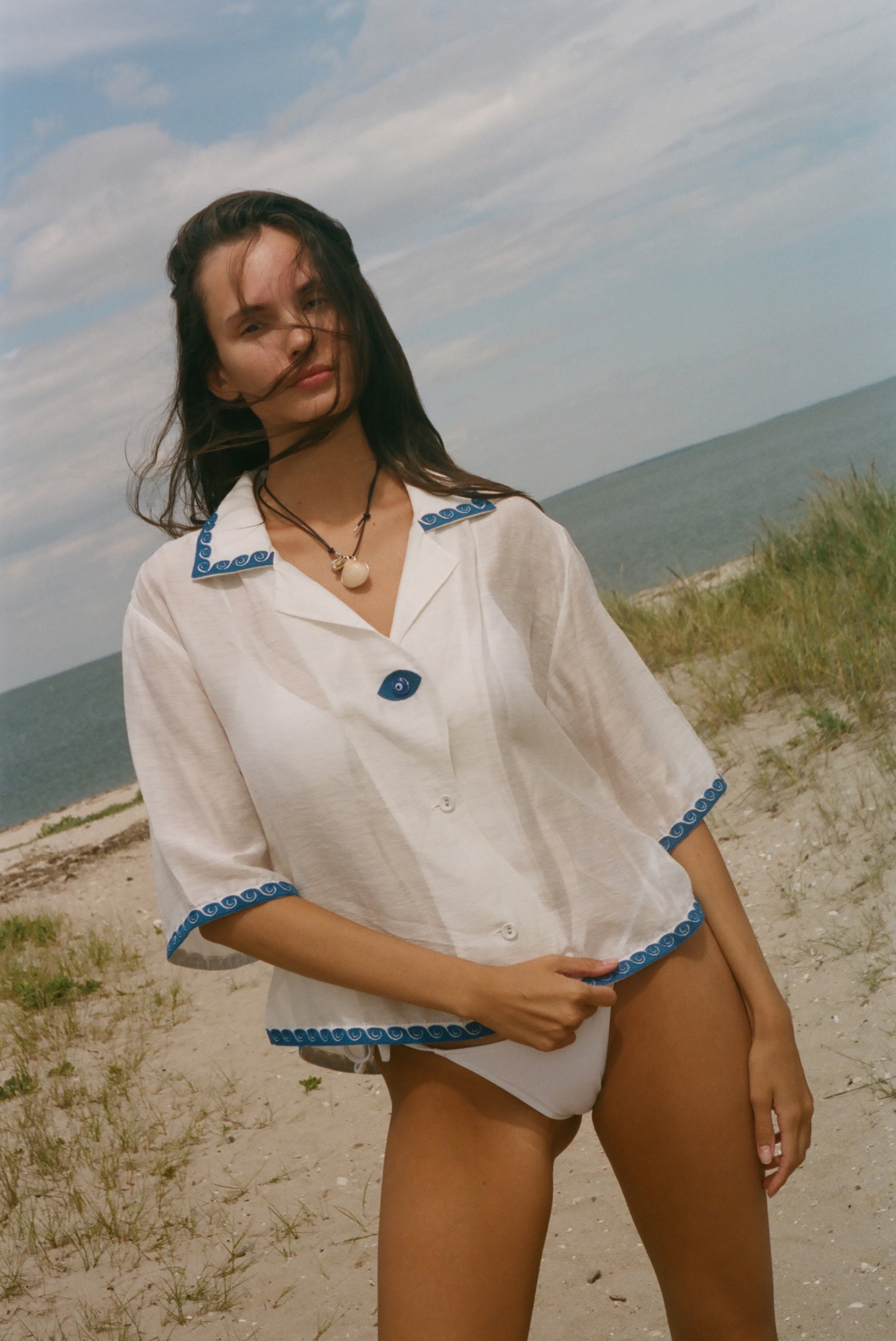 A woman stands on a sandy beach wearing a white short-sleeve button-front garment with blue trim along the collar, cuffs, and hem.