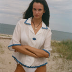 A woman standing on a sandy beach wearing a white short-sleeve collared shirt with blue trim and white bikini bottoms, with the ocean and cloudy sky in the background.