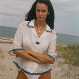 A woman standing on a sandy beach wearing a white short-sleeve collared shirt with blue trim and white bikini bottoms, with the ocean and cloudy sky in the background.