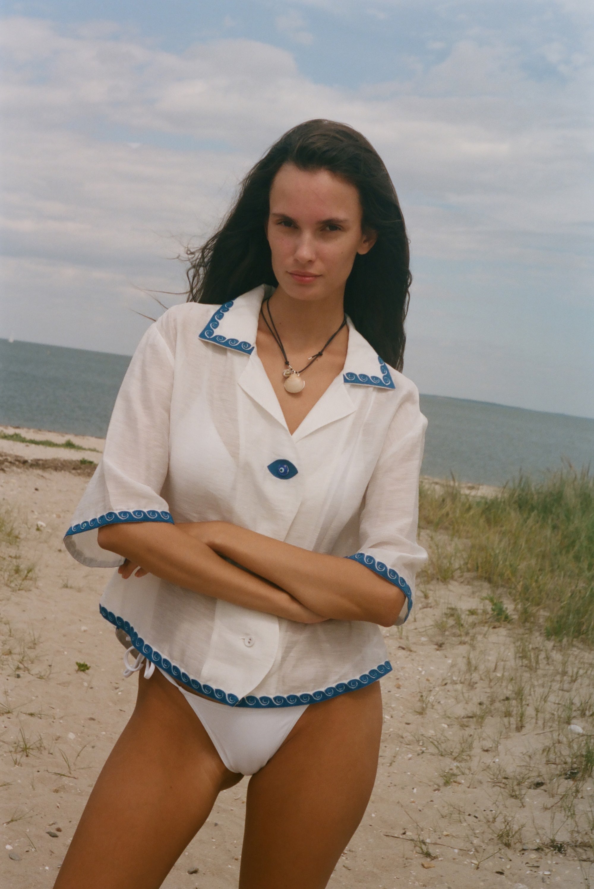 A woman standing on a sandy beach wearing a white short-sleeve collared shirt with blue trim and white bikini bottoms, with the ocean and cloudy sky in the background.