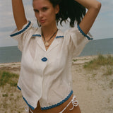 A woman posing on a sandy beach wearing a white short-sleeve button-up shirt with blue trim and matching white bikini bottoms.