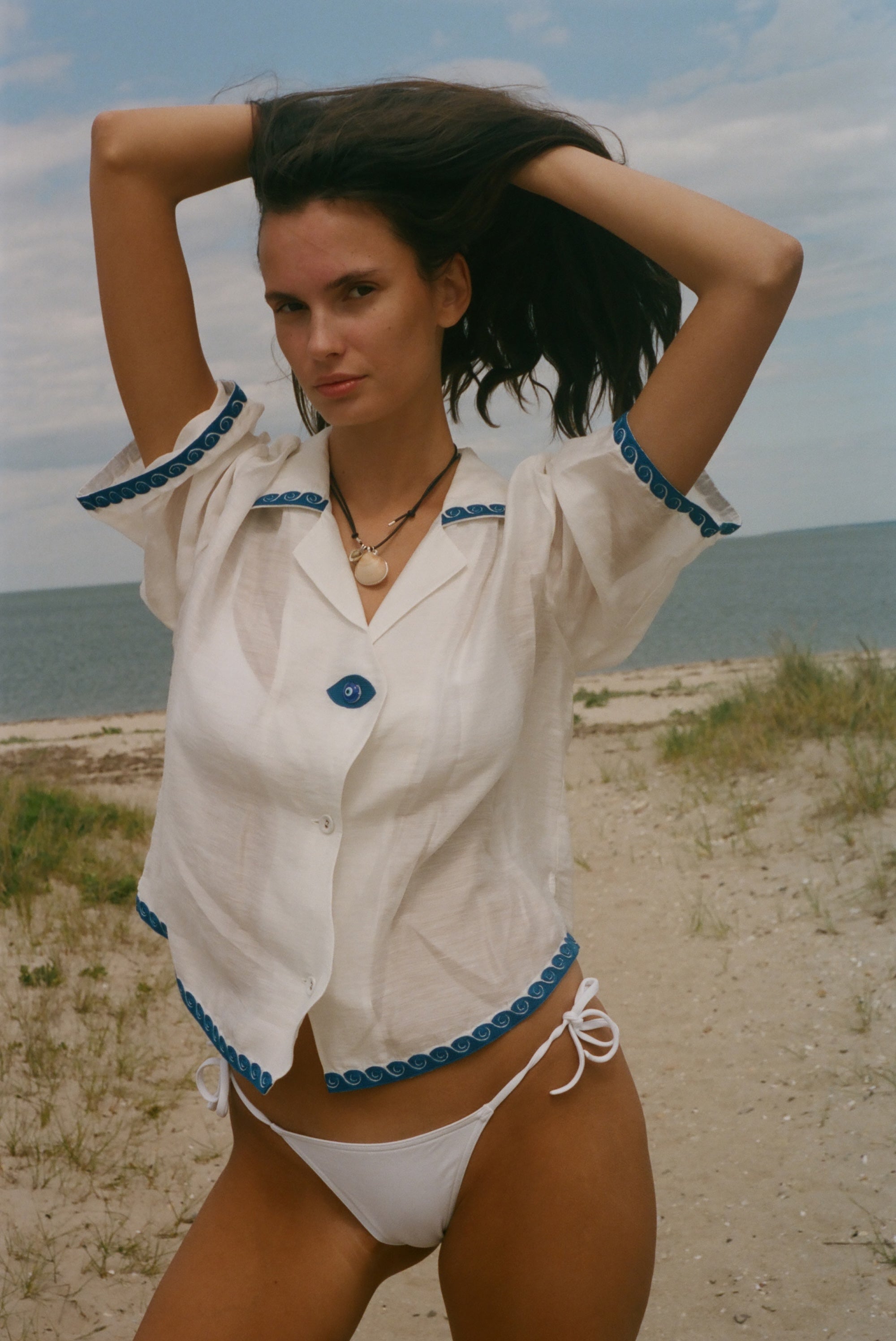 A woman posing on a sandy beach wearing a white short-sleeve button-up shirt with blue trim and matching white bikini bottoms.