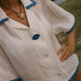 A close-up of a woman wearing a white short-sleeve button-front shirt with blue scalloped trim and a blue button, photographed outdoors near water and grass.