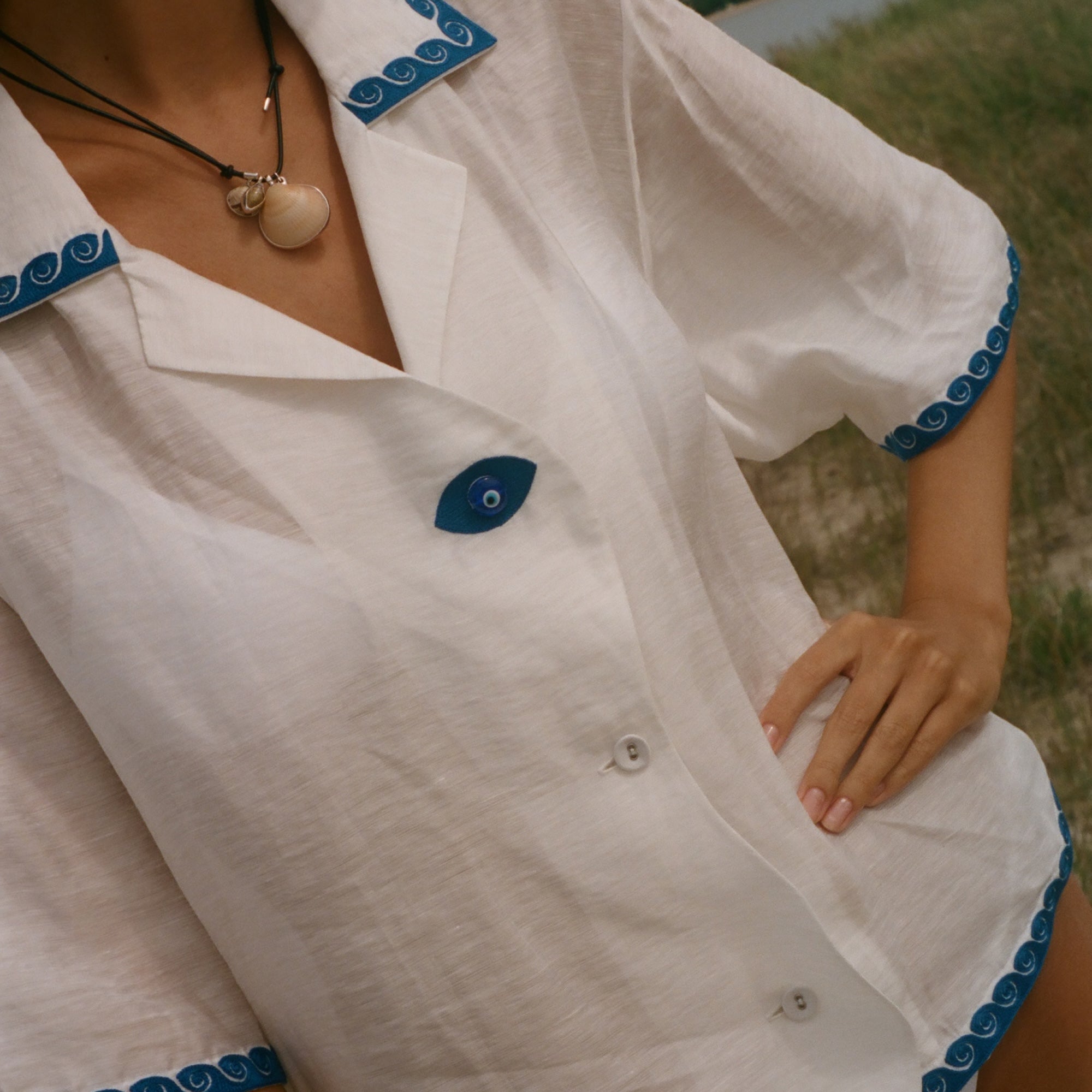 A close-up of a woman wearing a white short-sleeve button-front shirt with blue scalloped trim and a blue button, photographed outdoors near water and grass.