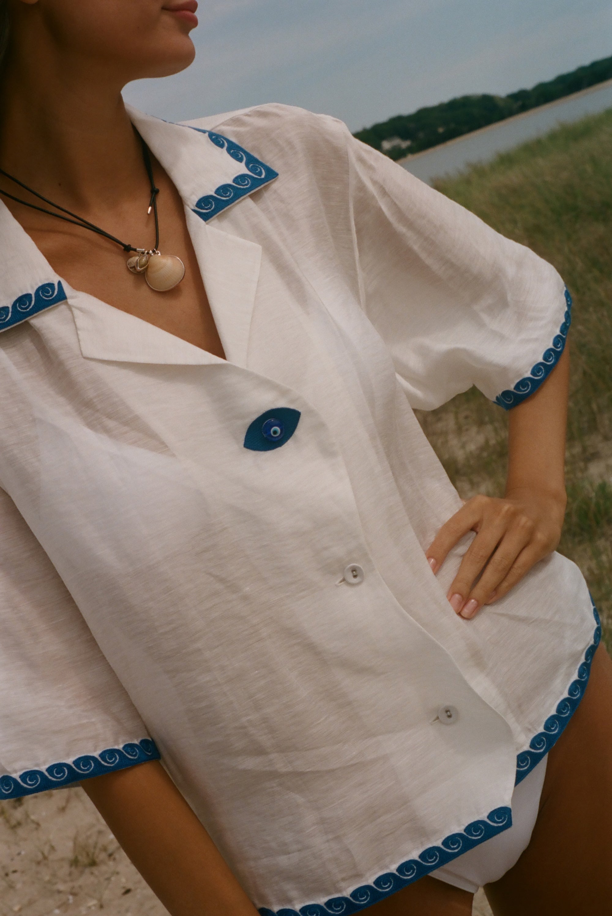 A close-up of a woman wearing a white short-sleeve button-front shirt with blue scalloped trim and a blue button, photographed outdoors near water and grass.