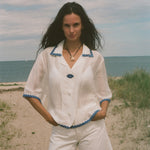 A woman stands on a sandy beach wearing a white short-sleeve button-front top with blue trim and matching white shorts.