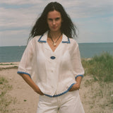 A woman stands on a sandy beach wearing a white short-sleeve button-front top with blue trim and matching white shorts.