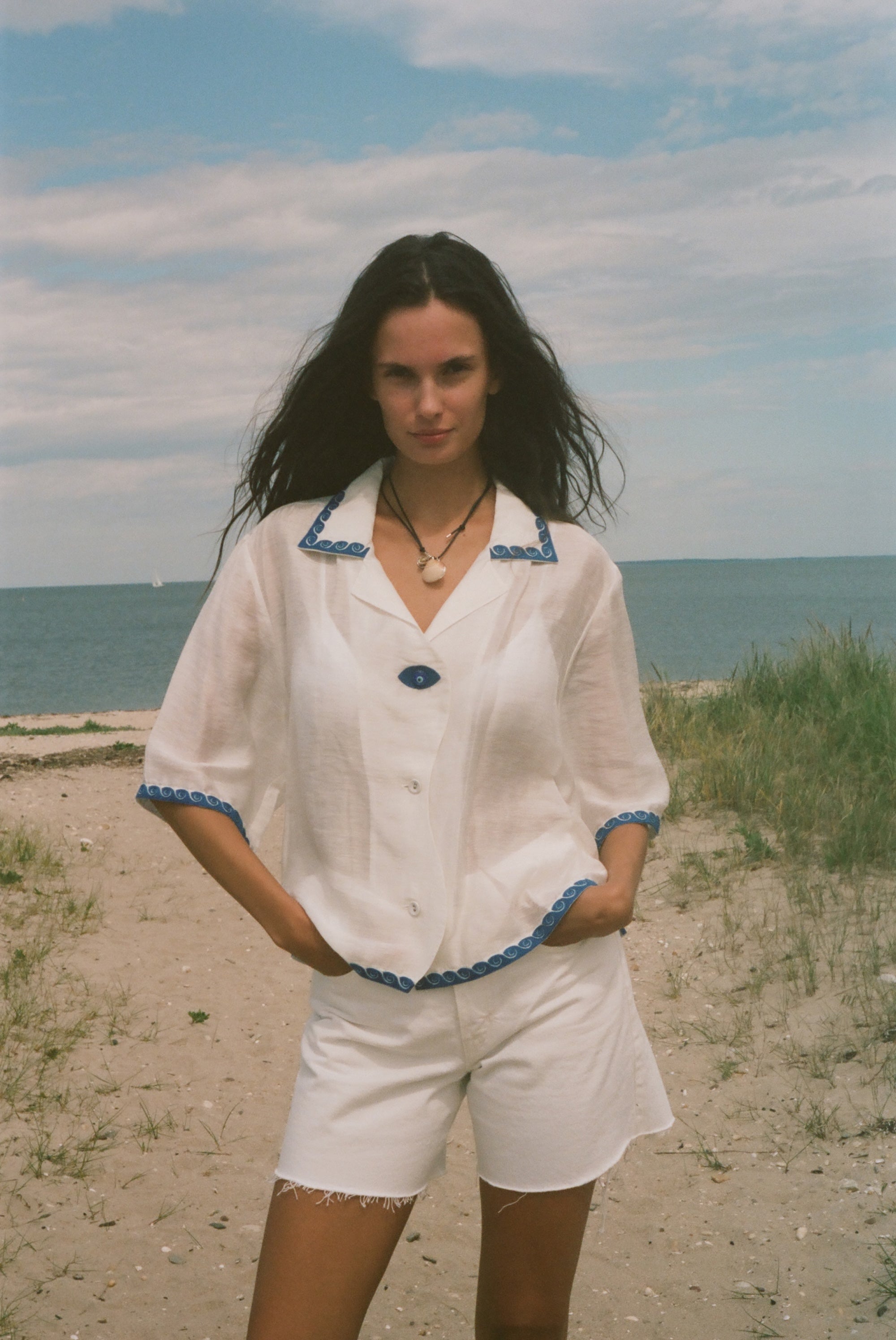 A woman stands on a sandy beach wearing a white short-sleeve button-front top with blue trim and matching white shorts.
