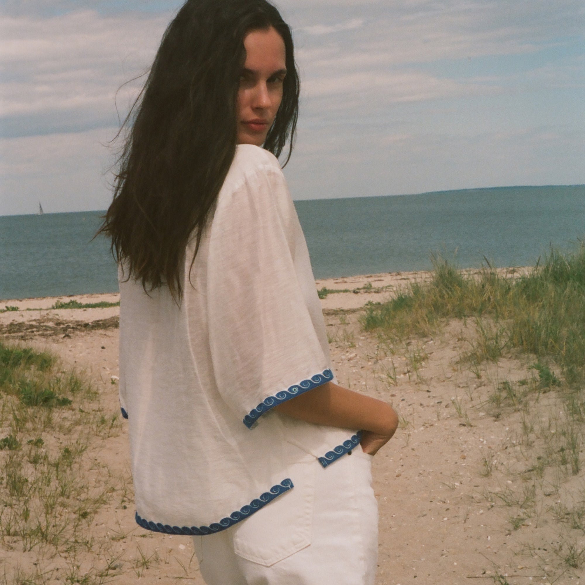 A woman standing on a sandy beach dune wearing a white short-sleeve top and white shorts with blue trim, looking back over her shoulder toward the camera.