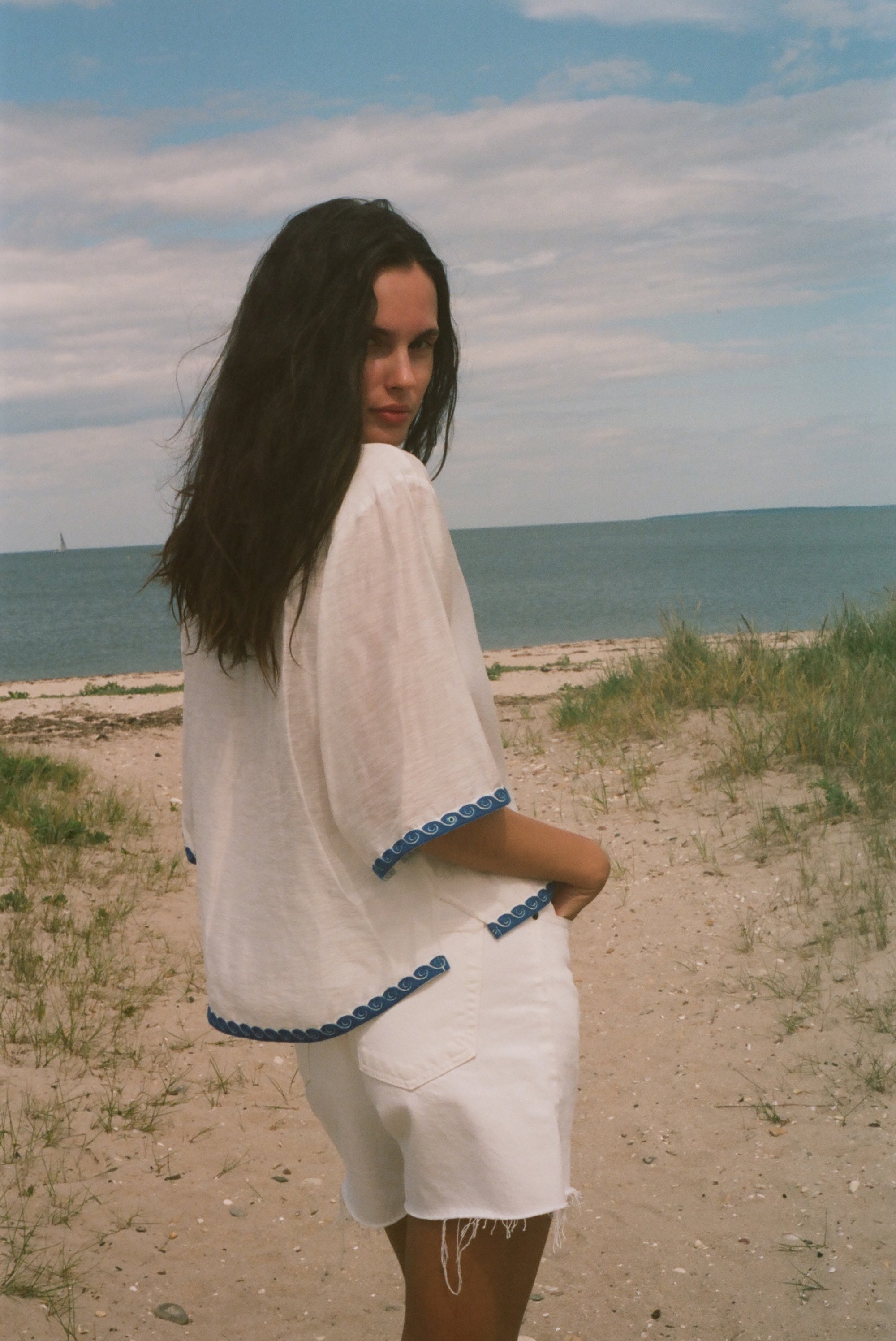 A woman standing on a sandy beach dune wearing a white short-sleeve top and white shorts with blue trim, looking back over her shoulder toward the camera.