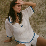 A woman sitting on sandy ground outdoors, wearing a white short-sleeve button-front top and matching shorts with blue trim details.