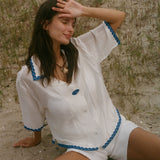 A woman sitting on sandy ground outdoors, wearing a white short-sleeve button-front top and matching shorts with blue trim details.