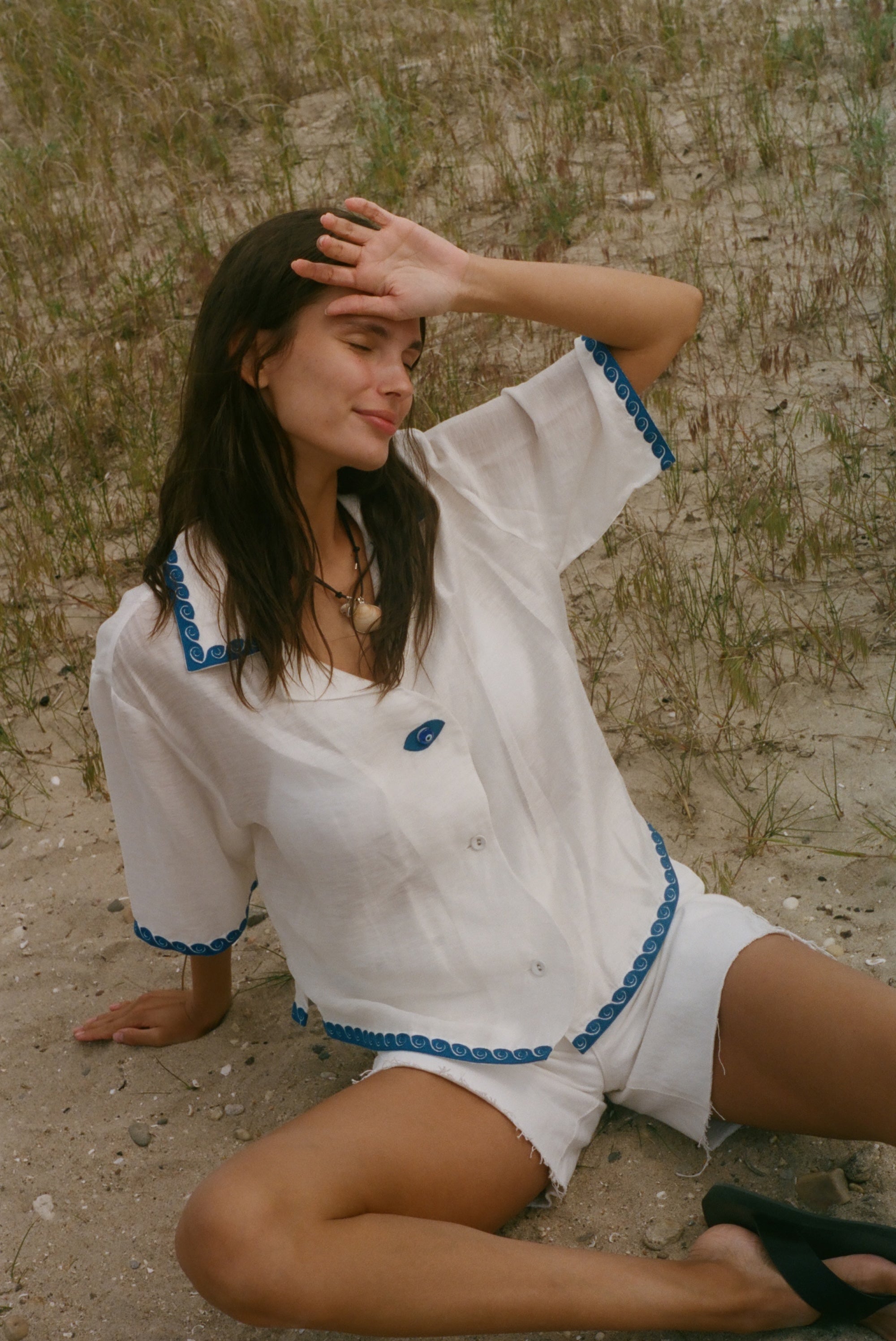A woman sitting on sandy ground outdoors, wearing a white short-sleeve button-front top and matching shorts with blue trim details.