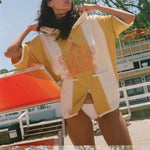 A woman wearing a yellow and white oversized beach poncho with colorful embroidery on the front, standing outdoors near a beach chair and trees.
