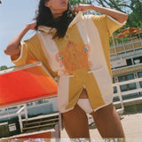 A woman wearing a yellow and white oversized beach poncho with colorful embroidery on the front, standing outdoors near a beach chair and trees.
