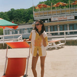 A woman wearing a yellow and white patterned short-sleeve shirt and matching shorts stands on a sandy beach next to an orange and white beach chair, with a building and trees in the background.