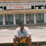 A man sits barefoot on concrete steps in front of a building labeled 'Sunset Beach' and 'Hotel, Restaurant & Bar'. He is wearing a white and light blue short-sleeve shirt with yellow shorts.
