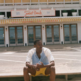 A man sits barefoot on concrete steps in front of a building labeled 'Sunset Beach' and 'Hotel, Restaurant & Bar'. He is wearing a white and light blue short-sleeve shirt with yellow shorts.