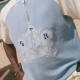 A close-up of a person wearing a light blue and off-white raglan quarter-zip t-shirt with a white embroidered mermaid graphic on the front, set against a beach background.
