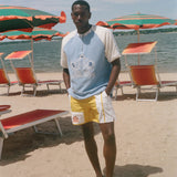 A man stands on a sandy beach wearing a light blue and white raglan t-shirt with a graphic print, yellow and white swim trunks, and standing barefoot in front of beach umbrellas and lounge chairs.