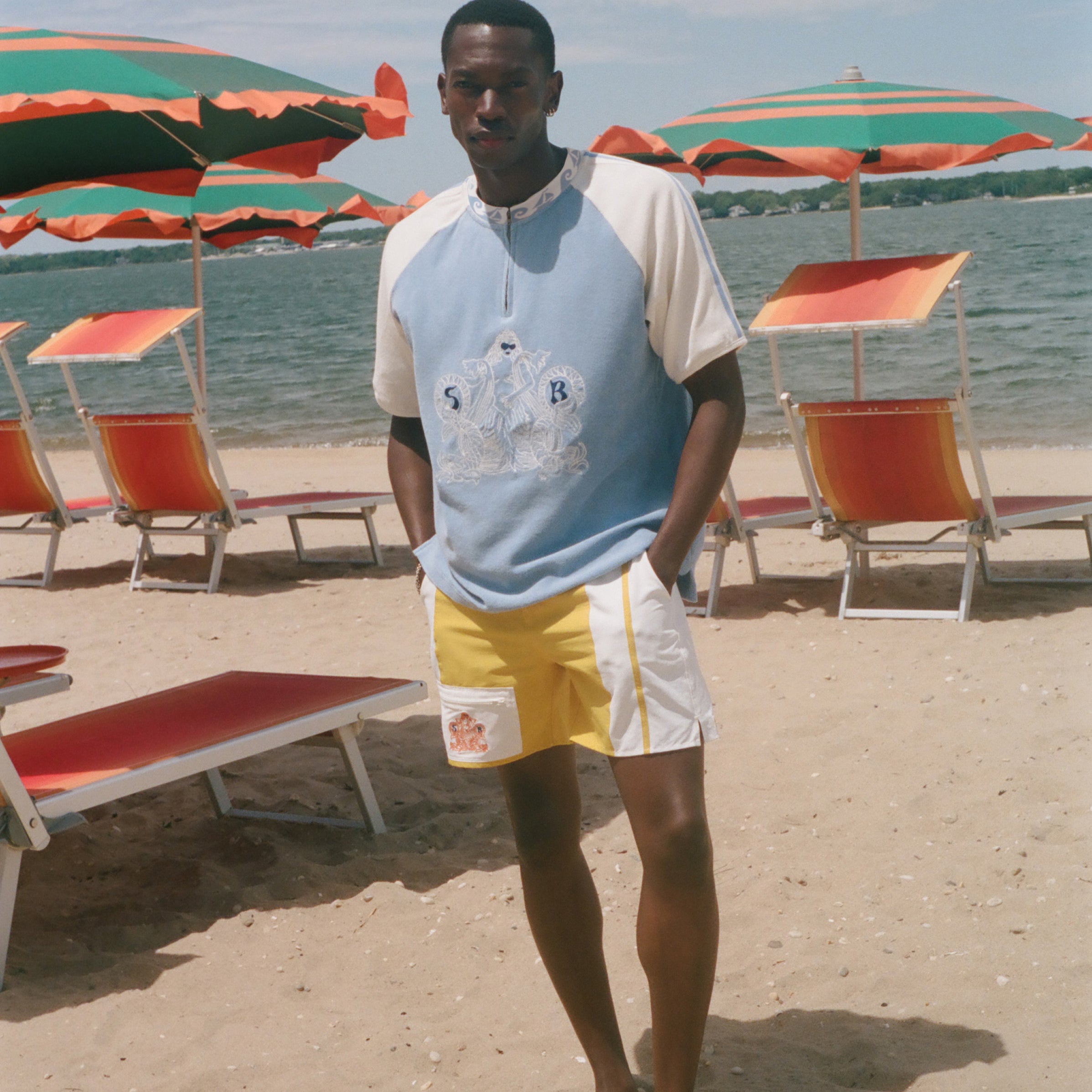A man stands on a sandy beach wearing a light blue and white raglan t-shirt with a graphic print, yellow and white swim trunks, and standing barefoot in front of beach umbrellas and lounge chairs.