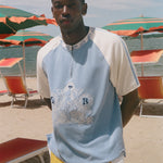 A man standing on a sandy beach wearing a light blue and off-white raglan t-shirt with a graphic print on the front, paired with yellow and white shorts. Beach umbrellas and lounge chairs are visible in the background.