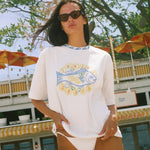 A woman wearing an oversized white graphic t-shirt with a blue and yellow fish design and the text 'SUNSET BEACH', photographed outdoors in front of a restaurant and trees.