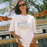 A woman wearing an oversized white graphic t-shirt with a blue and yellow fish design and the text 'SUNSET BEACH', photographed outdoors in front of a restaurant and trees.