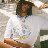 A woman wearing a white graphic t-shirt with a blue and yellow fish design, a straw hat, and sunglasses, sitting outdoors on a wooden bench.