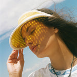 A woman outdoors wearing a yellow sun visor with a geometric dot pattern, with sunlight casting a matching pattern of shadows across her face. She is wearing a white t-shirt and a necklace.