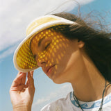 A woman outdoors wearing a yellow sun visor with a geometric dot pattern, with sunlight casting a matching pattern of shadows across her face. She is wearing a white t-shirt and a necklace.