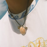 A close-up of a person wearing a dark cord necklace with a peach-colored shell pendant and small silver-toned accents, paired with a white graphic t-shirt.