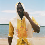 A man wearing a yellow and off-white hooded zip-up jacket with a large orange graphic on the front, standing on a sandy beach with water and sky in the background.