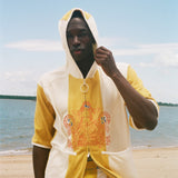 A man wearing a yellow and off-white hooded zip-up jacket with a large orange graphic on the front, standing on a sandy beach with water and sky in the background.