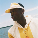 A man wearing a yellow and off-white hooded sweatshirt with a large graphic on the front, a matching yellow visor, and a black cap, standing on a beach with the ocean and blue sky in the background.
