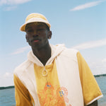 A man wearing a yellow and off-white hooded short-sleeve top with a graphic print stands on a beach with water and sky in the background.