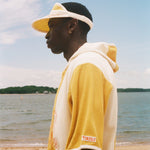 A man in profile wearing a yellow and off-white color-blocked hoodie and matching visor, standing on a sandy beach with water and sky in the background. 