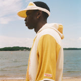 A man in profile wearing a yellow and off-white color-blocked hoodie and matching visor, standing on a sandy beach with water and sky in the background. 