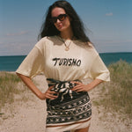 Woman standing on a beach wearing a 'Turismo' t-shirt and patterned skirt.