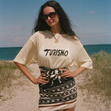 Woman standing on a beach wearing a 'Turismo' t-shirt and patterned skirt.