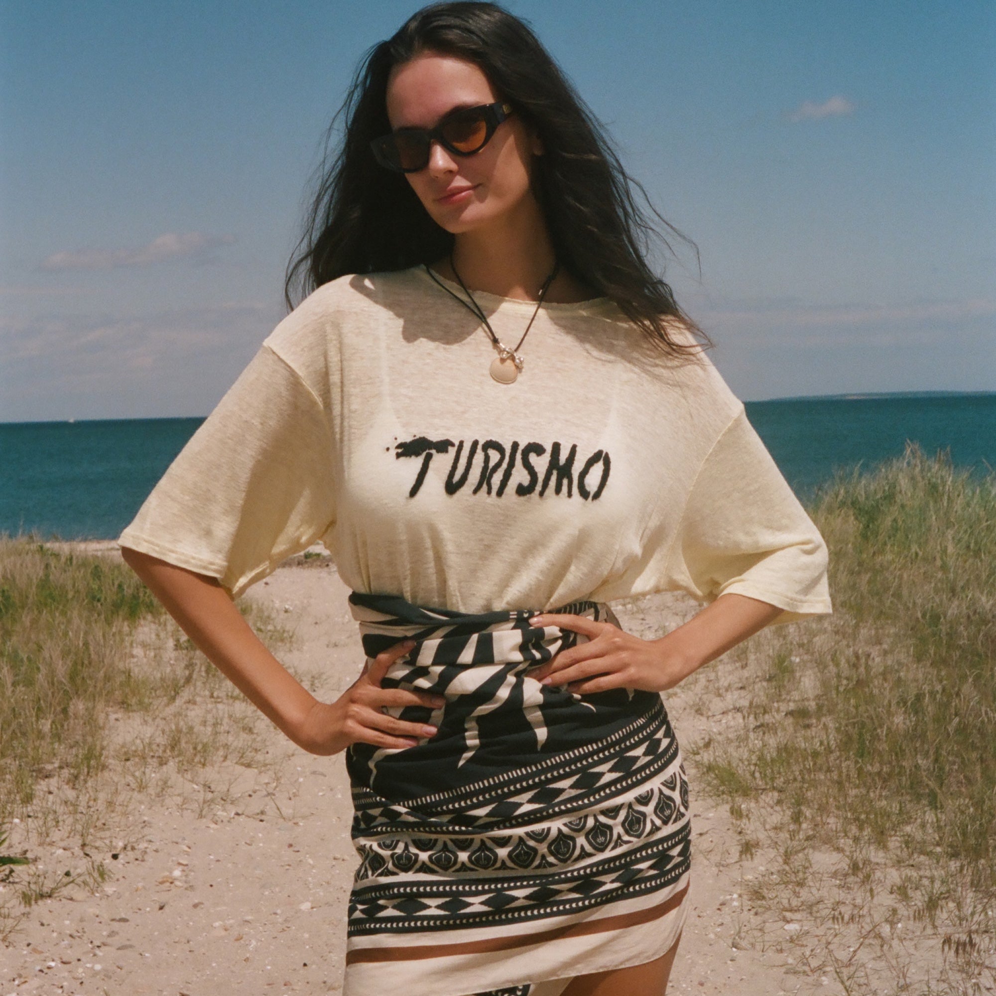 Woman standing on a beach wearing a 'Turismo' t-shirt and patterned skirt.