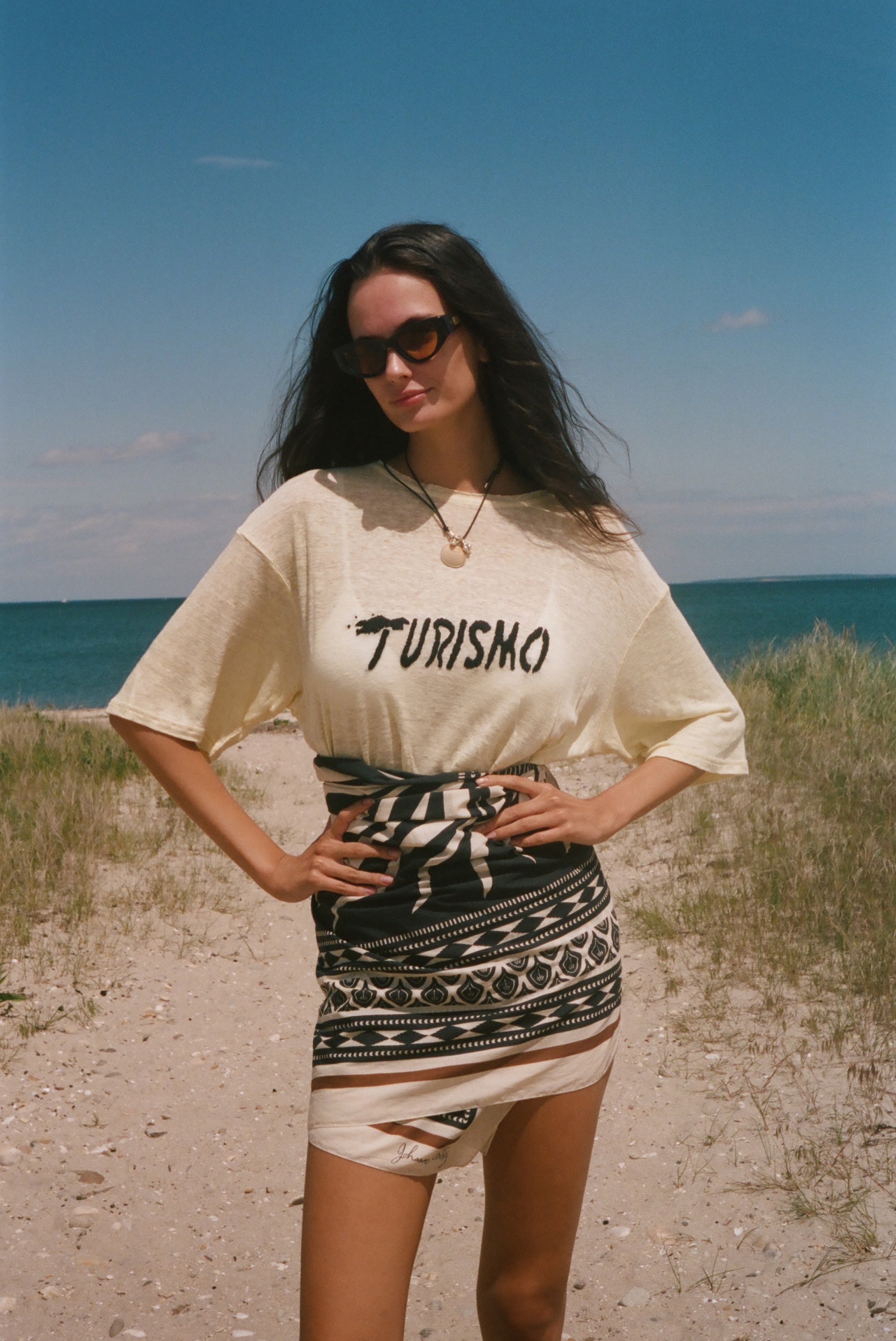 Woman standing on a beach wearing a 'Turismo' t-shirt and patterned skirt.