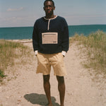 A man stands barefoot on a sandy path with beach grass and the ocean under a blue sky, wearing a dark navy long-sleeve sweater with a beige chest pocket and yellow shorts.