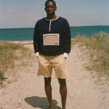 A man stands barefoot on a sandy path with beach grass and the ocean under a blue sky, wearing a dark navy long-sleeve sweater with a beige chest pocket and yellow shorts.