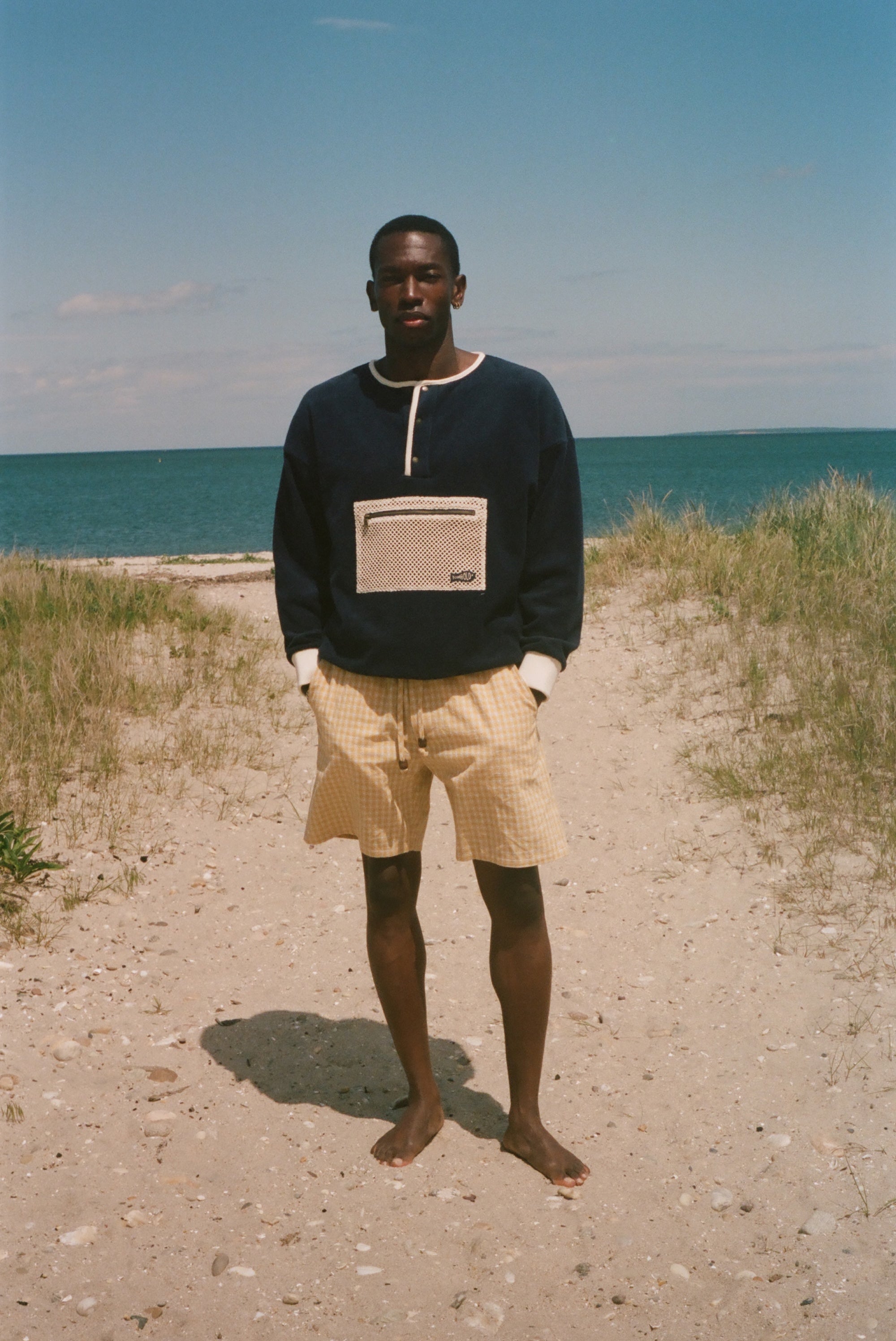 A man stands barefoot on a sandy path with beach grass and the ocean under a blue sky, wearing a dark navy long-sleeve sweater with a beige chest pocket and yellow shorts.