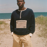 A man standing on a sandy beach wearing a navy blue long-sleeve henley sweatshirt with a beige mesh chest pocket and yellow gingham shorts, with the ocean and dunes in the background.
