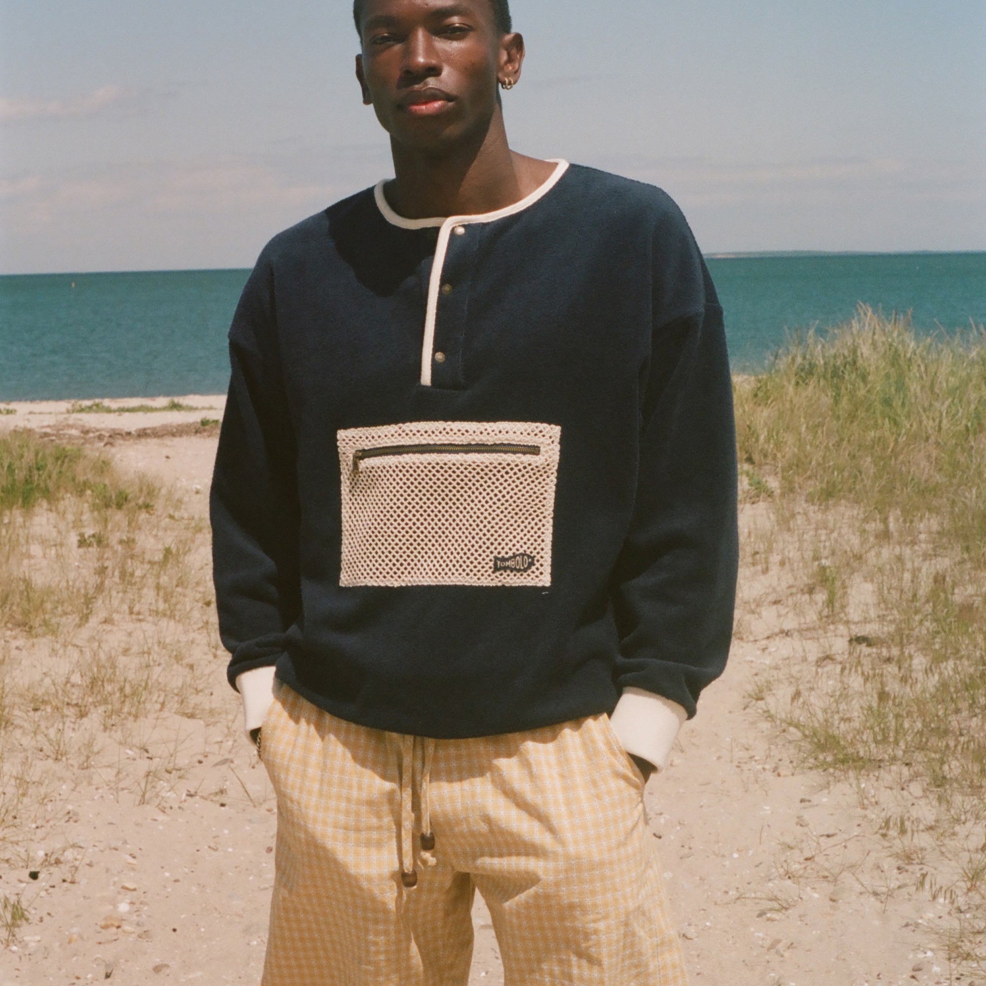A man standing on a sandy beach wearing a navy blue long-sleeve henley sweatshirt with a beige mesh chest pocket and yellow gingham shorts, with the ocean and dunes in the background.