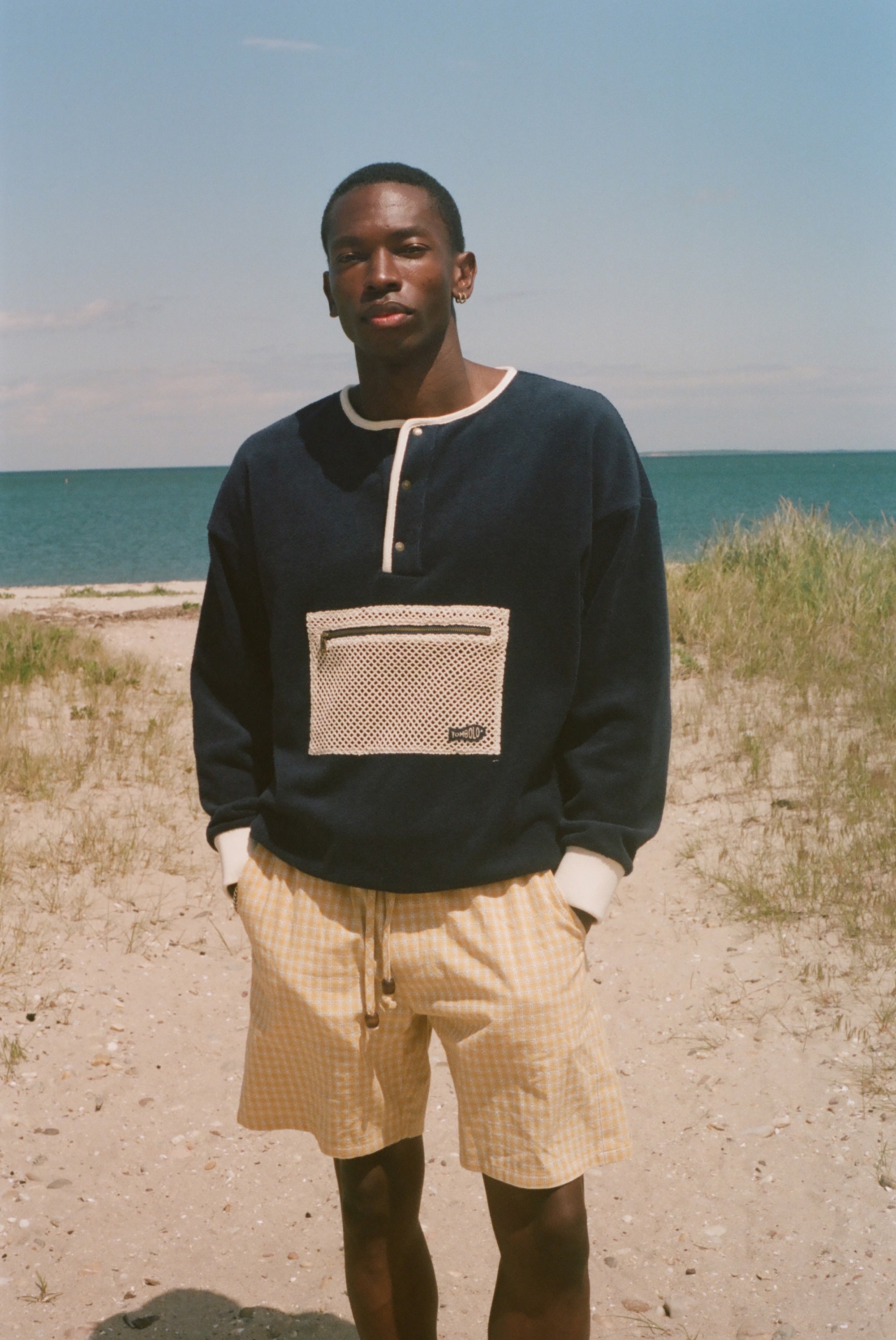 A man standing on a sandy beach wearing a navy blue long-sleeve henley sweatshirt with a beige mesh chest pocket and yellow gingham shorts, with the ocean and dunes in the background.