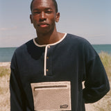 A man wearing a navy blue long-sleeve henley sweater with a beige mesh chest pocket, standing outdoors on a sandy beach with the ocean and sky in the background.