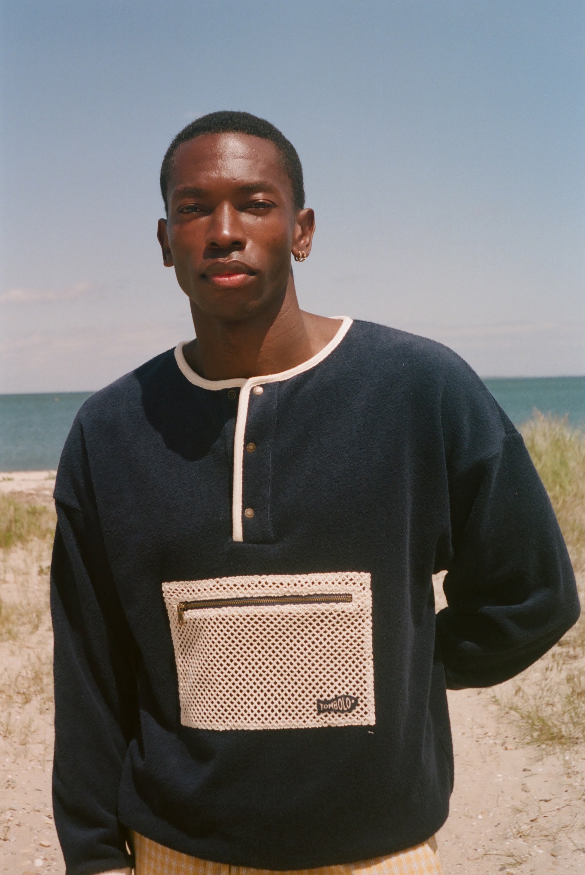 A man wearing a navy blue long-sleeve henley sweater with a beige mesh chest pocket, standing outdoors on a sandy beach with the ocean and sky in the background.